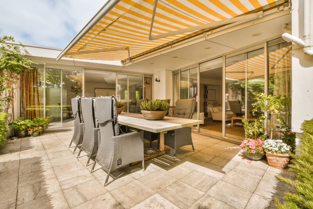 Patio with a table and chairs under a striped awning, enhancing outdoor living space with greenery and flowers, illustrating the benefits of patio covers for shade and aesthetic appeal.