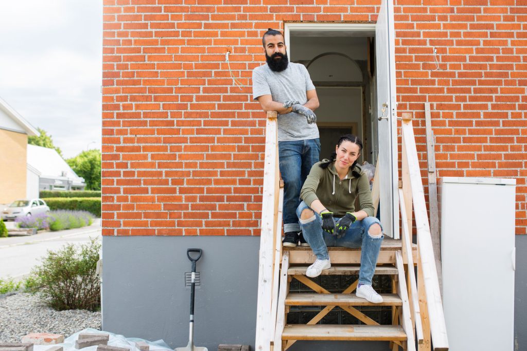Couple standing on wooden stairs outside a brick apartment, preparing for home renovation projects in spring.