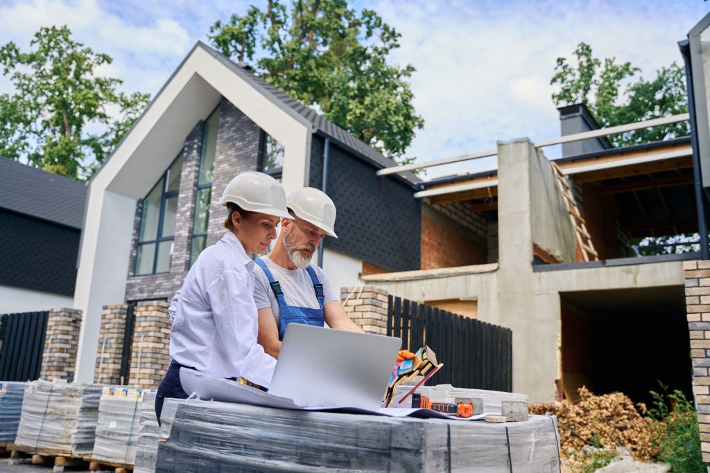 Construction professionals reviewing plans on a laptop at a remodeling site, surrounded by building materials and a partially constructed home, emphasizing personalized consultation and tailored strategies in home remodeling.
