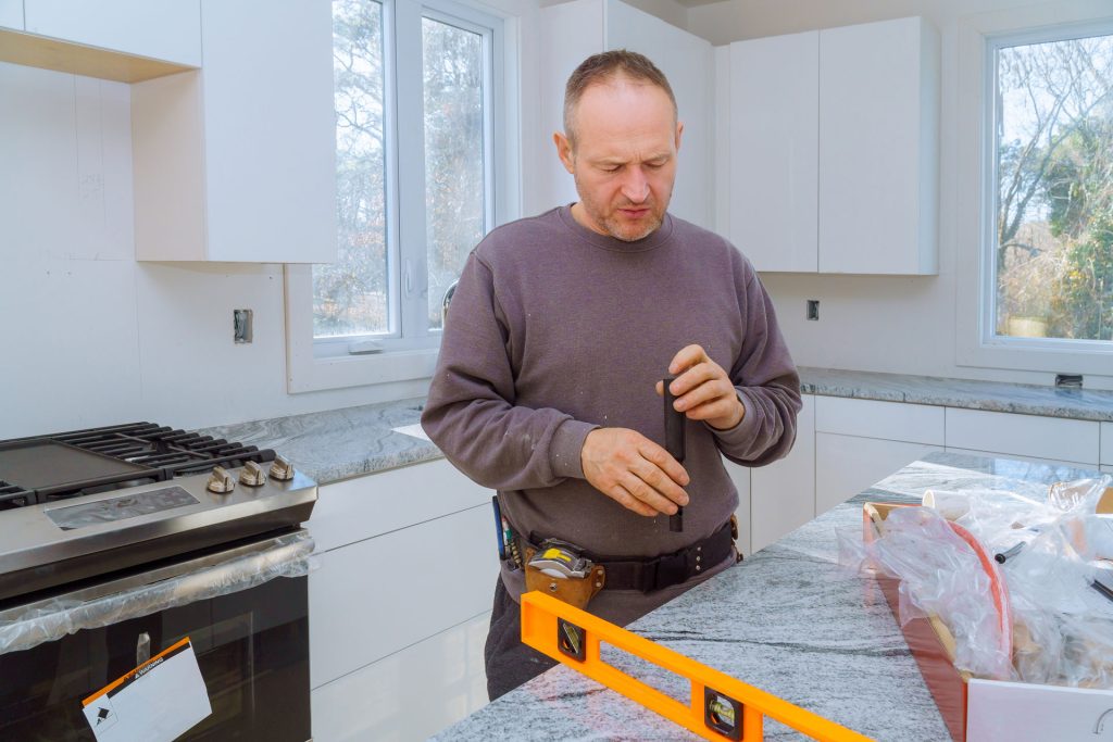 Plumber assembling and installing faucet in modern kitchen with granite countertops and white cabinetry.