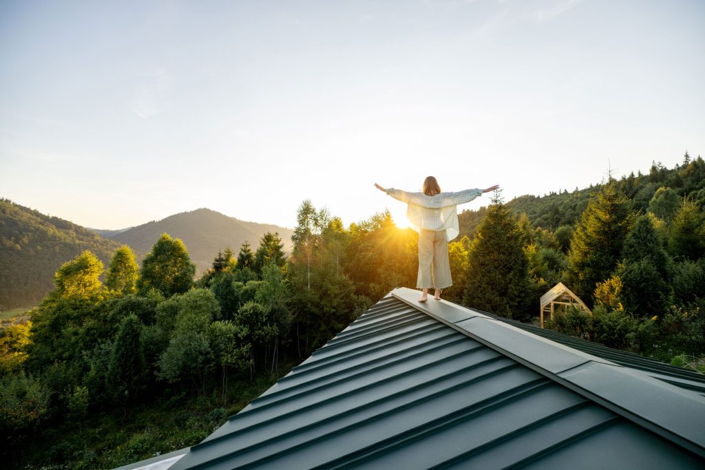 Woman standing on a metal rooftop with arms outstretched, overlooking a mountainous landscape at sunrise, symbolizing the importance of roof maintenance in Redding's climate.