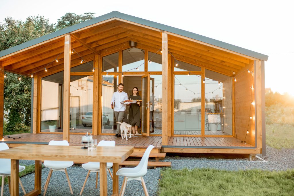 Friends enjoying a casual dinner near a modern accessory dwelling unit, featuring a wooden deck and string lights, with a dog and table set for dining.