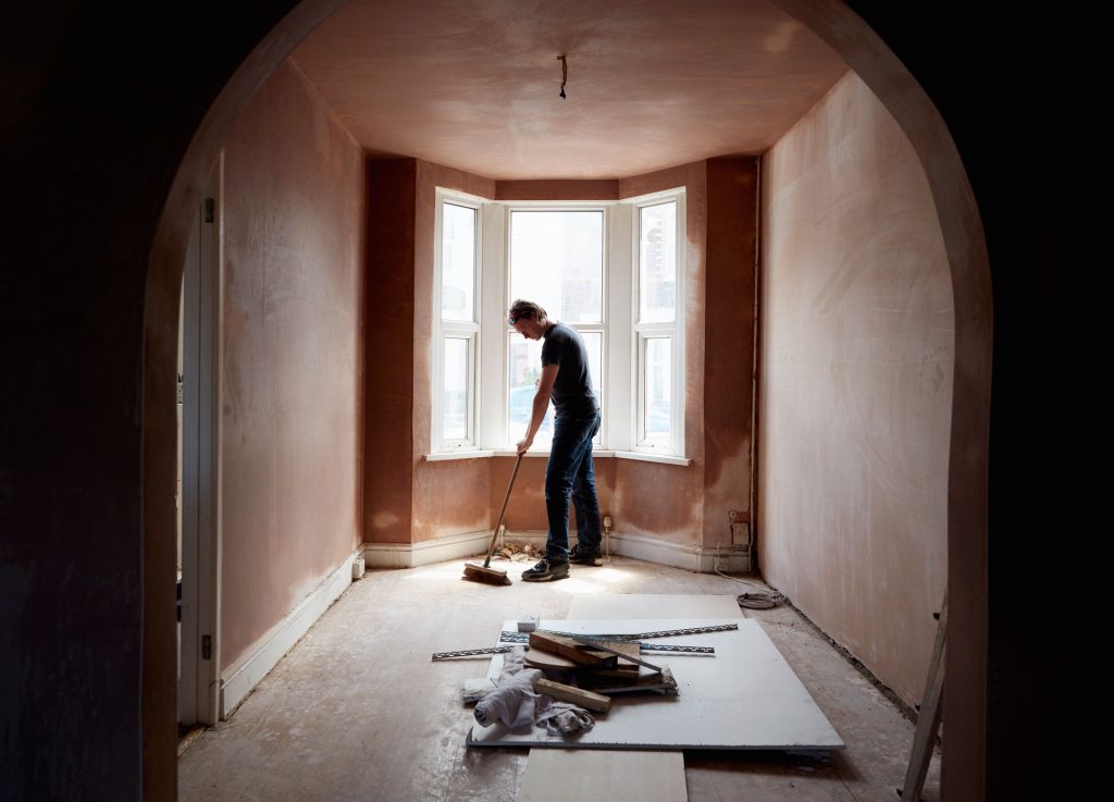 Builder sweeping and tidying up in a renovated room with natural light from bay windows, showcasing a clean, updated space ready for finishing touches.